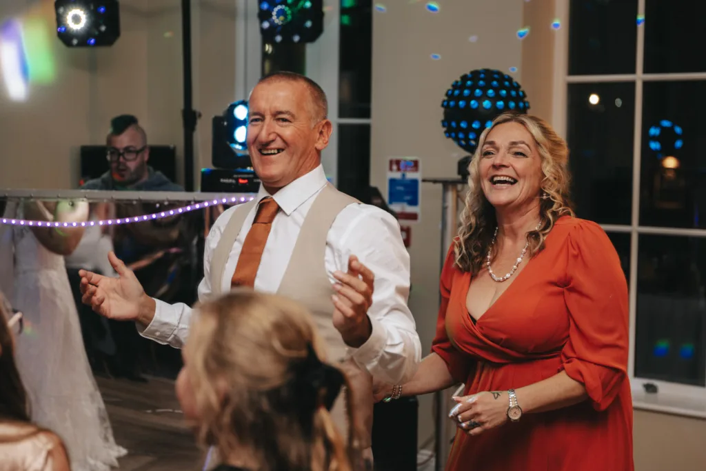 A man and a woman, both smiling, are dancing together at an indoor event at the Stallingborough Grange Hotel. The man wears a white shirt, beige vest, and orange tie. The woman is in an autumn-hued dress with pearl jewelry. Other people are visible in the background, and colorful lights illuminate the room. © Aimee Lince Photography - Wedding photographer in Lincolnshire, Yorkshire & Nottinghamshire