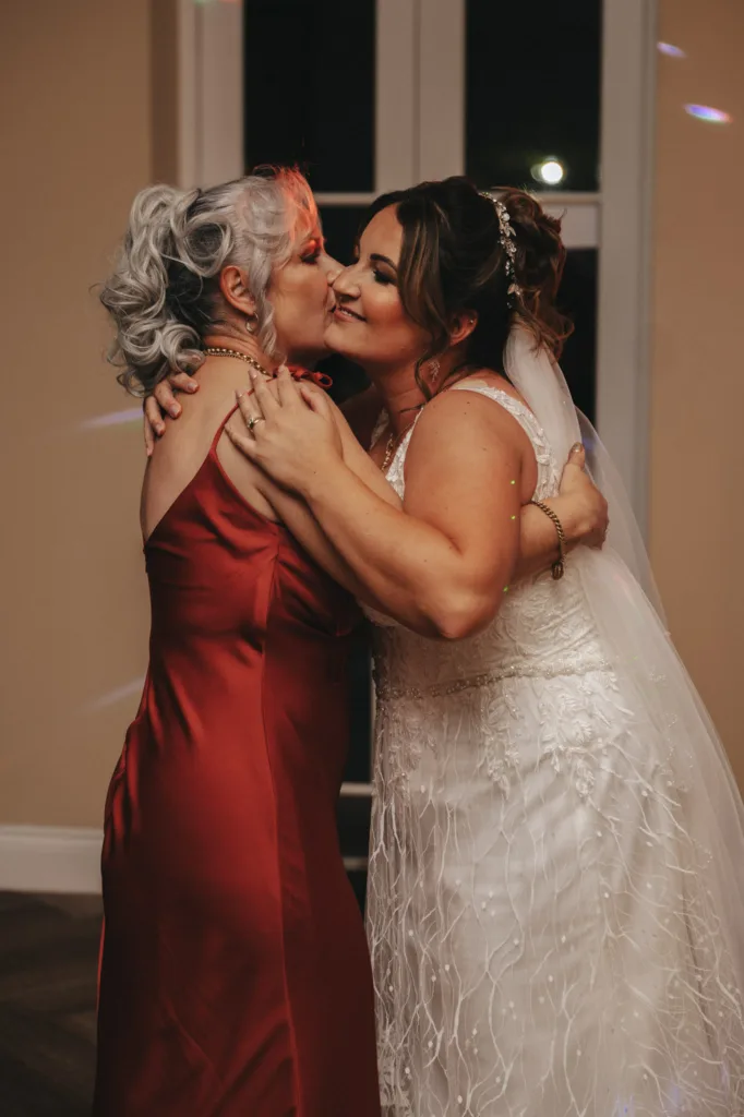A bride in a lace wedding dress and tiara smiles while embracing an older woman in a red dress during her enchanting autumn wedding. Her hair is styled in an updo as they share a joyful moment in the warmly lit room with large windows at Stallingborough Grange Hotel. © Aimee Lince Photography - Wedding photographer in Lincolnshire, Yorkshire & Nottinghamshire