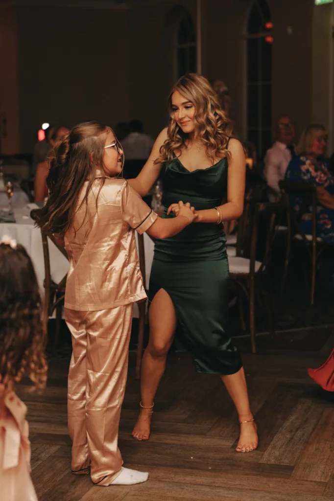 At an autumn wedding indoors at Stallingborough Grange Hotel, two people dance gracefully. One dazzles in a shiny, light pink outfit with glasses, the other in a sleeveless green dress with a slit. They hold hands and smile fondly at each other while guests sit in the softly lit background. © Aimee Lince Photography - Wedding photographer in Lincolnshire, Yorkshire & Nottinghamshire