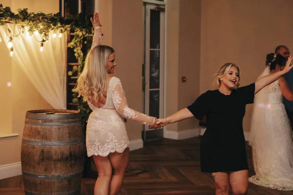 Two women dance joyfully, holding hands, in an autumn-lit room. The woman on the left wears a white lace dress and raises her arm triumphantly. Her companion in a black dress smiles warmly, extending her arm out. Nearby, a rustic wooden barrel complements the cozy atmosphere of this Stallingborough Grange Hotel celebration. © Aimee Lince Photography - Wedding photographer in Lincolnshire, Yorkshire & Nottinghamshire