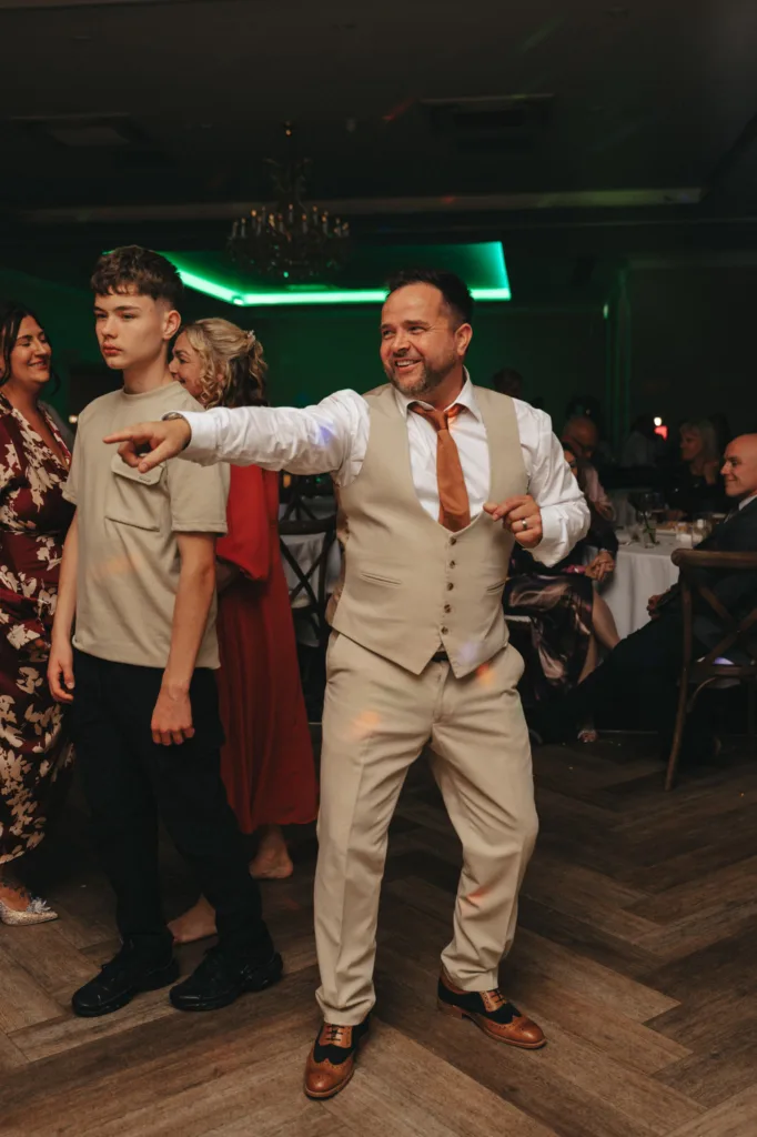 A cheerful man in a beige three-piece suit and brown tie dances enthusiastically, pointing with one hand on a wooden floor in a dimly lit room with green accent lighting. Nearby, at an autumn wedding at Stallingborough Grange Hotel, a teenage boy in a beige shirt and black pants stands. Guests sit at round tables in the background. © Aimee Lince Photography - Wedding photographer in Lincolnshire, Yorkshire & Nottinghamshire