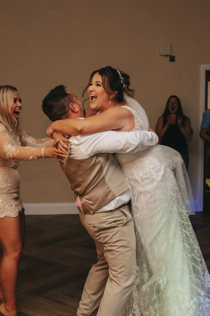 At the Stallingborough Grange Hotel, a groom in a tan suit lifts the bride, who is wearing a white lace gown, as they both laugh joyfully. A woman in a light dress stands nearby, cheering and clapping amidst the warm hues of an autumn wedding. Another person in the background also claps with enthusiasm. © Aimee Lince Photography - Wedding photographer in Lincolnshire, Yorkshire & Nottinghamshire