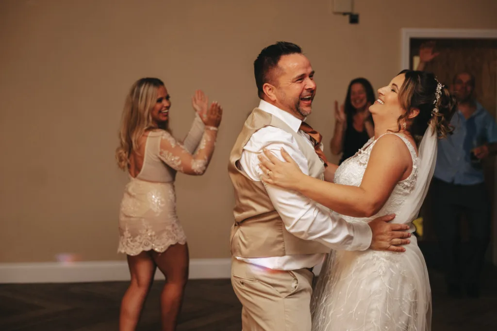 At Stallingborough Grange, a bride in a white dress and veil dances closely with her groom in a beige vest and white shirt. The couple smiles joyfully, embracing the warm autumn theme. In the background, two women and a man clap along, one wearing a light pink dress applauding enthusiastically. © Aimee Lince Photography - Wedding photographer in Lincolnshire, Yorkshire & Nottinghamshire