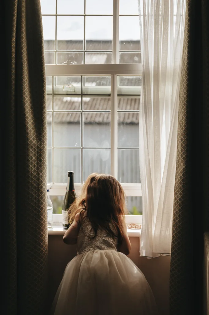 A young girl with long hair, wearing a white dress, leans on a windowsill, gazing outside. The window has a grid pattern, and soft autumn light filters in through sheer curtains. A bottle and small plate rest on the sill, while the background reveals a building with a corrugated roof. © Aimee Lince Photography - Wedding photographer in Lincolnshire, Yorkshire & Nottinghamshire