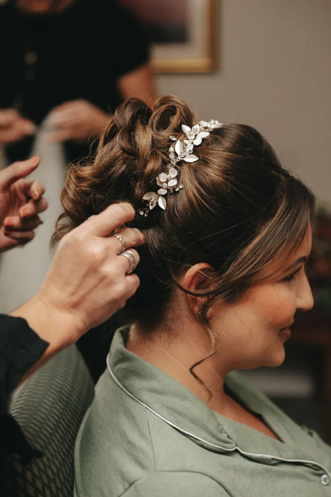 A person with curly, brown hair styled in an elegant updo adorned with a delicate floral hairpiece. A hand is adjusting the hair, reflecting an autumn theme. Dressed in a sage green outfit, they sit on a patterned chair at Stallingborough Grange, where soft, warm lighting creates a relaxed atmosphere. © Aimee Lince Photography - Wedding photographer in Lincolnshire, Yorkshire & Nottinghamshire