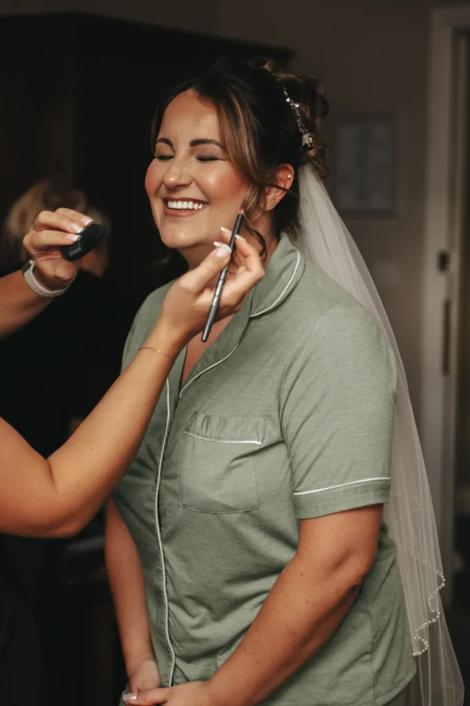 A smiling woman in a green pajama set is having makeup applied, her veil framing elegant hair styled for an autumn theme. Two hands work skillfully, one holding a brush to her cheek and the other dabbing with a sponge. The softly blurred background at Stallingborough Grange Hotel highlights her joyful expression. © Aimee Lince Photography - Wedding photographer in Lincolnshire, Yorkshire & Nottinghamshire