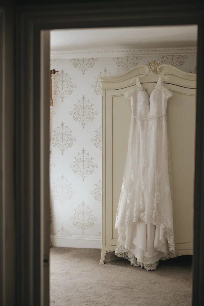 A white lace wedding dress hangs elegantly on the doors of a vintage wardrobe. The room, perfect for an autumn wedding, features ornate floral-patterned wallpaper in soft gold tones. The beige carpet and gentle natural light create a serene and romantic atmosphere. © Aimee Lince Photography - Wedding photographer in Lincolnshire, Yorkshire & Nottinghamshire
