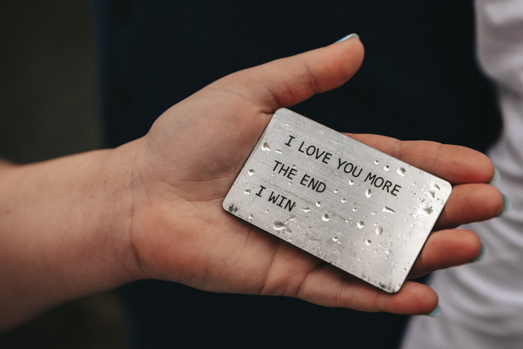 A hand holds a weathered metal card stamped with the text "I LOVE YOU MORE — THE END — I WIN," reminiscent of heartfelt notes exchanged in People's Park. Raindrops dot its surface, and the person's nails are painted light blue. © Aimee Lince Photography