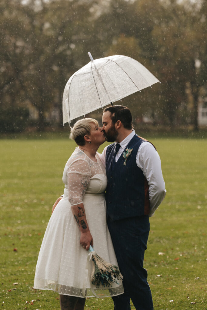 In a romantic wedding shoot at People's Park, a newlywed couple kisses under a clear umbrella in a grassy field as rain falls gently. The bride, in a white dress with sheer sleeves, holds a bouquet, while the groom dons a blue vest and tie. Soft, overcast light and blurred trees enhance the intimate atmosphere. © Aimee Lince Photography