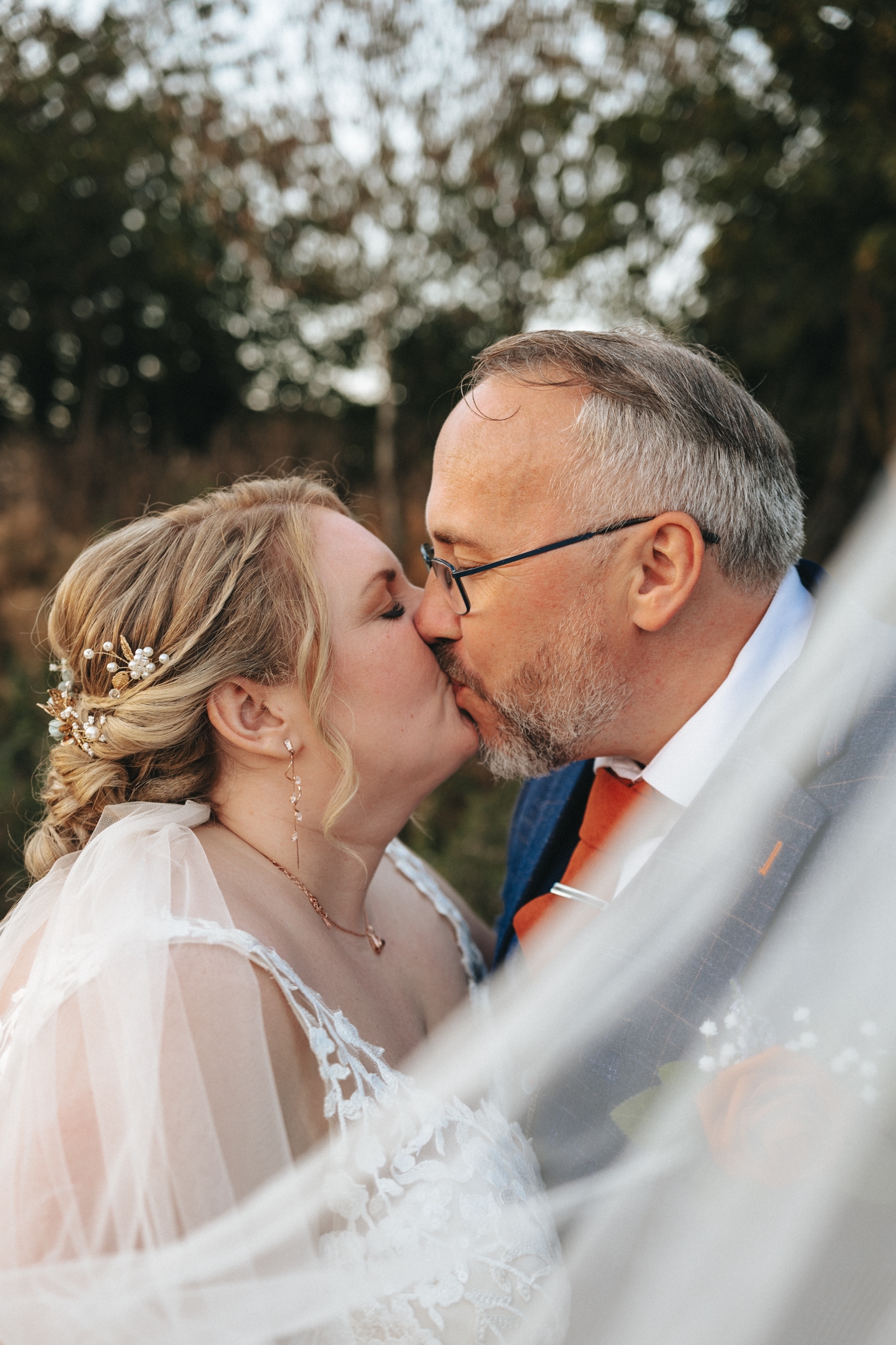 Kirsty and John share a tender kiss, framed by flowing white veil fabric at their sunny September wedding. The bride's lace dress and floral hairpiece complement the groom's suit and orange tie, as they stand outdoors in Cleethorpes, surrounded by blurred trees, capturing a romantic moment. © Aimee Lince Photography