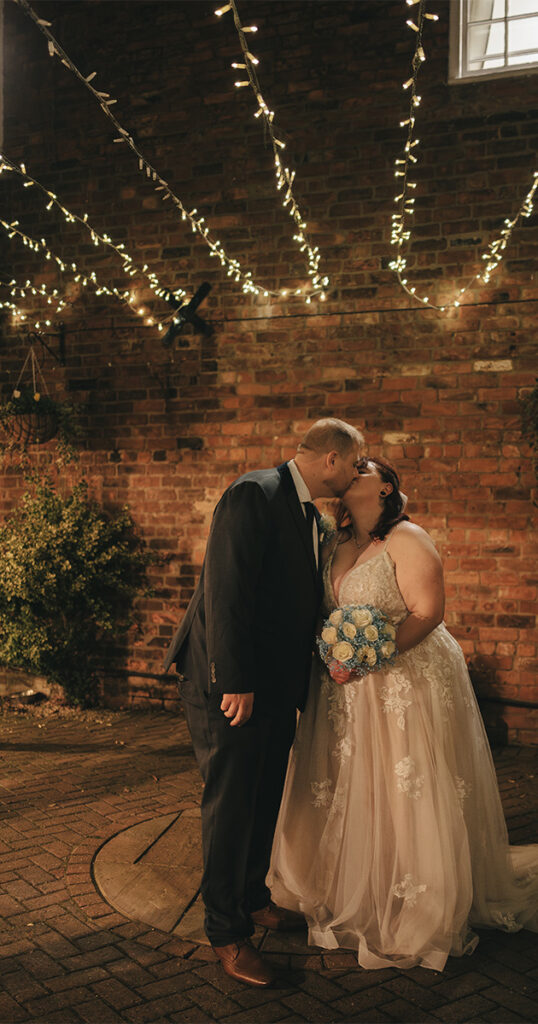 Alicia & Reece share a tender kiss under warm fairy lights against a brick wall. The bride, in a lace dress with pastel blooms, and the groom, in a dark suit, are framed by greenery. Their Wonderland Wedding at Admiral Rodney exudes intimate romance. © Aimee Lince Photography