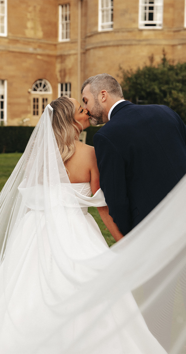 A bride in a white gown and veil kisses the groom, wearing a dark suit, outdoors. They're standing in front of a large, historic building with arched windows and brick walls; Rudding Park Spa. Her veil flows elegantly, partially covering them and creating a timeless and elegant atmosphere. © Aimee Lince Photography