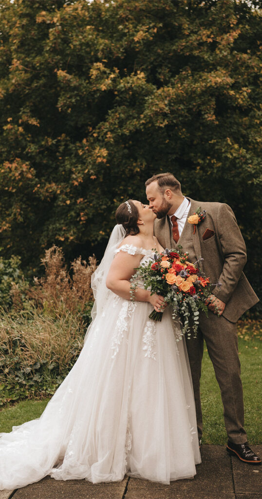 A bride in a white lace wedding dress and veil kisses a groom in a brown suit with a red tie during their autumnal themed wedding. The groom holds a bouquet of red, orange, and yellow flowers. They stand on a paved path with lush green trees at The Ashbourne Hotel, capturing Olivia & Lloyd's romantic outdoor setting. © Aimee Lince Photography