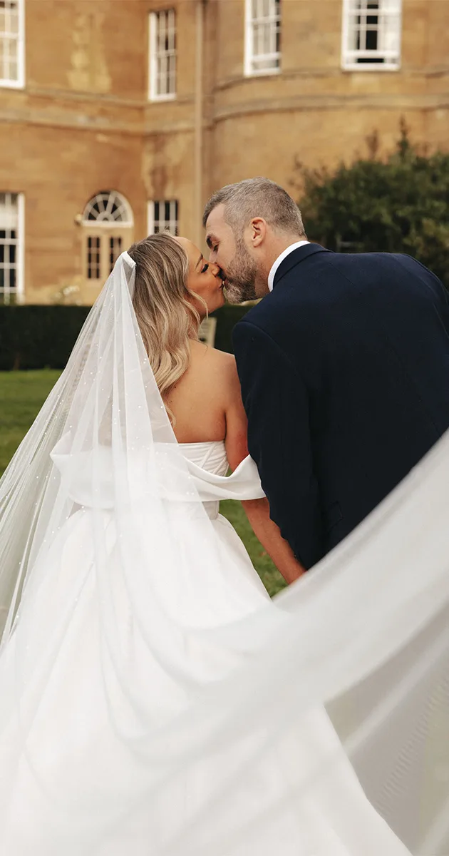 A bride and groom kiss in front of a historic stone building at Rudding Park. The bride, in an off-shoulder white gown and veil, faces away from the camera while the groom wears a dark suit. They hold hands, and the flowing veil captures the breeze, adding motion to this romantic setting. © Aimee Lince Photography - Wedding photographer in Lincolnshire, Yorkshire & Nottinghamshire