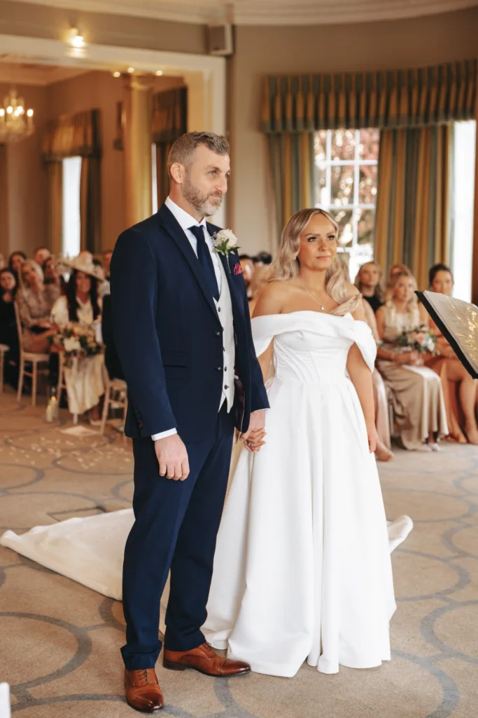 A couple stands holding hands in a wedding ceremony at Rudding Park in North Yorkshire. The groom dons a navy suit with a vest and tie, while the bride dazzles in an off-shoulder gown. Facing the officiant, guests seated against elegant drapes and chandeliers complete this Harrogate affair's charm. © Aimee Lince Photography - Wedding photographer in Lincolnshire, Yorkshire & Nottinghamshire