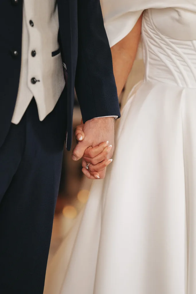 A couple holding hands at Rudding Park in Harrogate, dressed in formal wedding attire. The groom is in a dark suit with a white shirt and waistcoat, while the bride wears a white gown with a fitted bodice. Their interlocked hands stand out against the contrasting fabric of their outfits. © Aimee Lince Photography - Wedding photographer in Lincolnshire, Yorkshire & Nottinghamshire