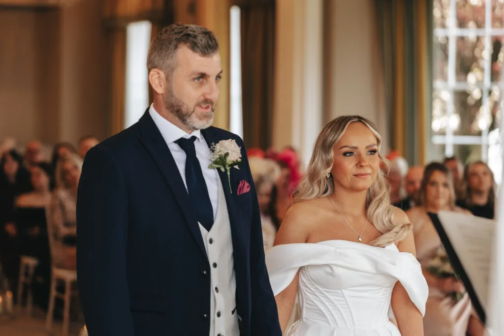 A couple stands elegantly in a formal setting at Rudding Park. The man wears a dark suit with a white vest and pink boutonnière, while the woman stuns in an off-shoulder white dress. Both have light hair, and guests are seated in the softly lit room with large windows overlooking Harrogate's charm. © Aimee Lince Photography - Wedding photographer in Lincolnshire, Yorkshire & Nottinghamshire