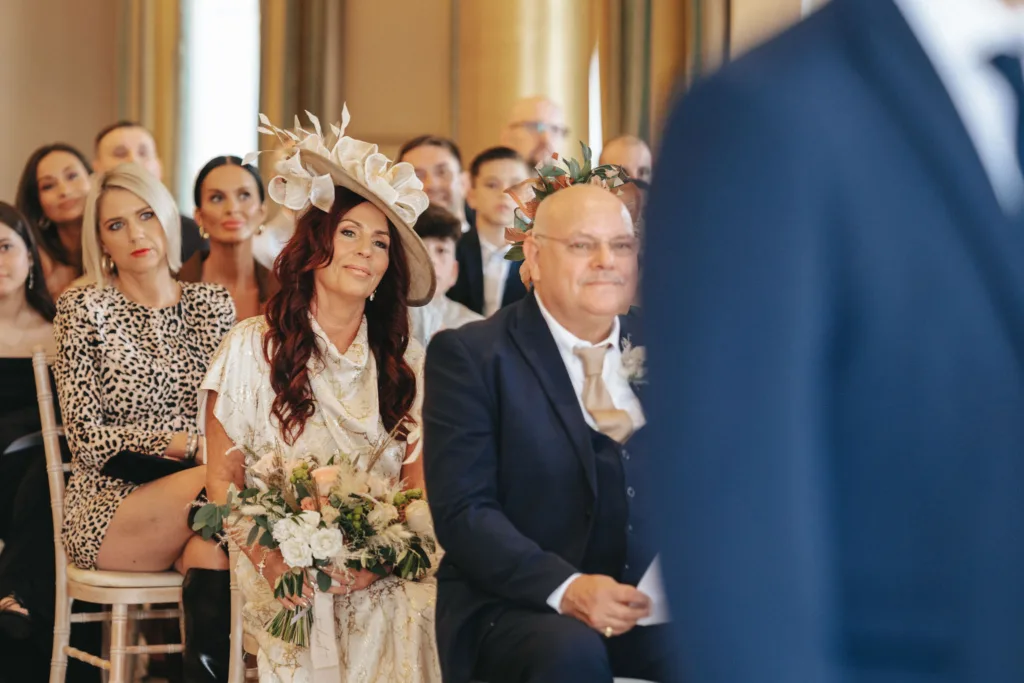 Guests seated at a wedding ceremony in the picturesque setting of Rudding Park, Harrogate. A woman with long red hair in a white floral dress and hat, holding a bouquet, sits beside a man in a dark suit, focusing on the speaker. Other attendees in formal wear sit closely behind them. © Aimee Lince Photography - Wedding photographer in Lincolnshire, Yorkshire & Nottinghamshire