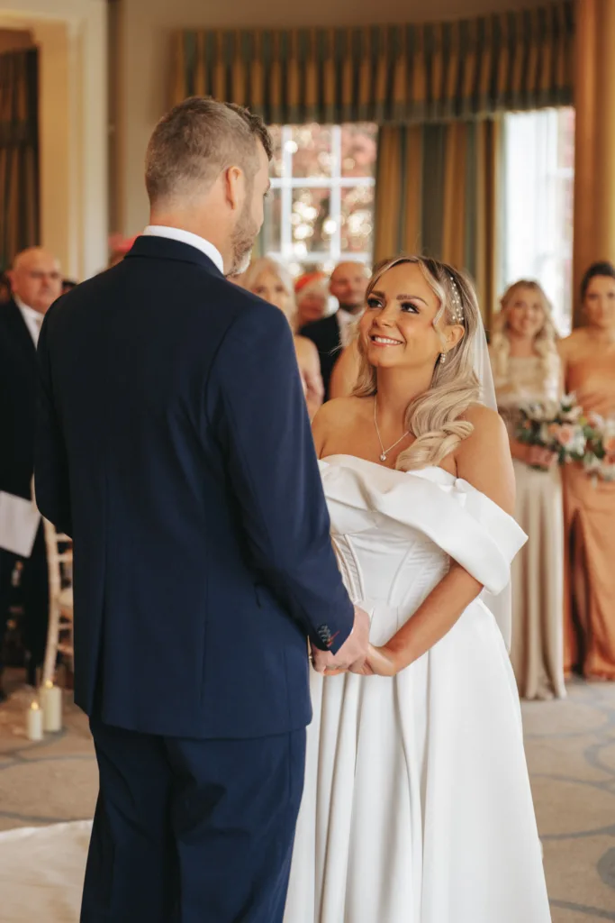 In a warmly lit room at Rudding Park in Harrogate, North Yorkshire, a bride in an off-the-shoulder white dress and veil smiles at her groom in a dark suit. Guests are seated in the background while bridesmaids in peach dresses stand with bouquets, witnessing the joyful moment. © Aimee Lince Photography - Wedding photographer in Lincolnshire, Yorkshire & Nottinghamshire