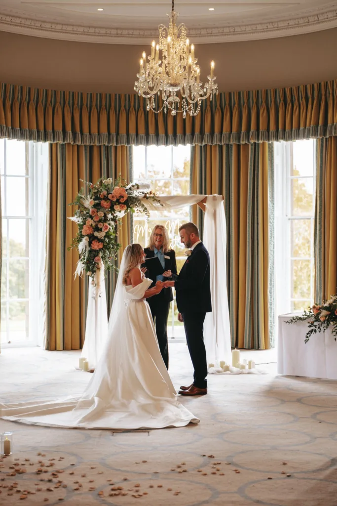 In a charming setting reminiscent of North Yorkshire's elegance, a bride in a white gown and groom in a dark suit hold hands beneath a floral arch with a neon sign. A chandelier glimmers above them, illuminating the room with large windows and gold curtains, while rose petals adorn the floor. © Aimee Lince Photography - Wedding photographer in Lincolnshire, Yorkshire & Nottinghamshire