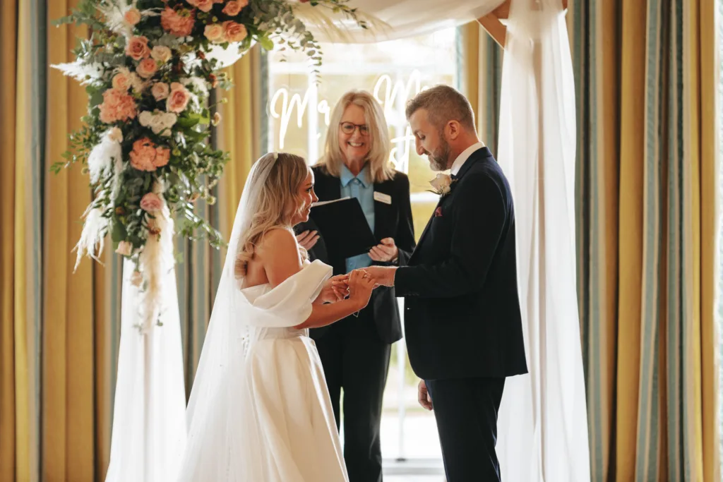 In the enchanting setting of Rudding Park, North Yorkshire, a beautiful wedding ceremony unfolds as the couple exchanges rings. The bride, wearing a white dress with off-the-shoulder sleeves, faces her groom in his dark suit beneath a floral arch adorned with white and pink flowers. © Aimee Lince Photography - Wedding photographer in Lincolnshire, Yorkshire & Nottinghamshire