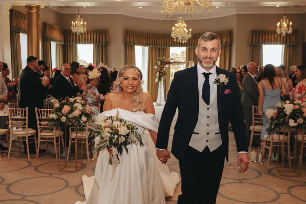 A bride and groom walk down the aisle at Rudding Park in Harrogate, smiling and holding hands. The bride wears an off-the-shoulder gown with a bouquet of white flowers. The groom is in a dark suit with a gray vest and black tie. Guests stand and clap in a hall adorned with chandeliers and large windows. © Aimee Lince Photography - Wedding photographer in Lincolnshire, Yorkshire & Nottinghamshire
