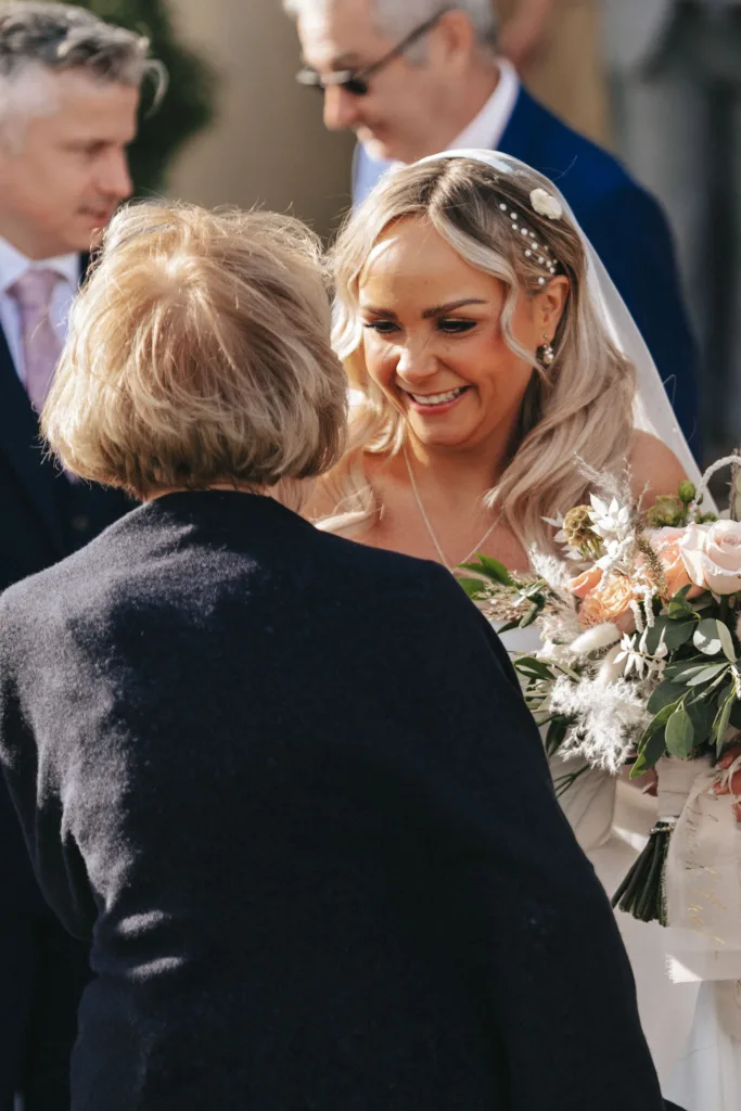 A bride with long blonde hair, adorned with pearls, smiles warmly while holding a bouquet of white and peach flowers at Rudding Park in Harrogate. She is wearing a white gown and veil. An older woman with short, light brown hair, wearing a dark coat, faces her. Two men in suits are in the background. © Aimee Lince Photography - Wedding photographer in Lincolnshire, Yorkshire & Nottinghamshire