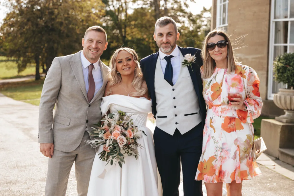 A wedding scene unfolds at Rudding Park, Harrogate, with four people beaming. The bride dazzles in an off-shoulder white dress, clutching a bouquet of pink and white flowers. The groom stands smartly in a dark suit with a gray vest, joined by two guests: one in a light gray suit and the other sporting a floral dress with sunglasses. © Aimee Lince Photography - Wedding photographer in Lincolnshire, Yorkshire & Nottinghamshire