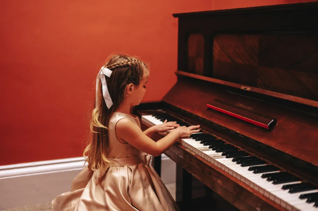 A young girl with long hair tied with a white ribbon is playing an upright piano at Rudding Park. She wears a satin pink dress, focused on the keys. The warm, solid orange wall creates a cozy atmosphere in this serene corner of North Yorkshire near Harrogate. © Aimee Lince Photography - Wedding photographer in Lincolnshire, Yorkshire & Nottinghamshire