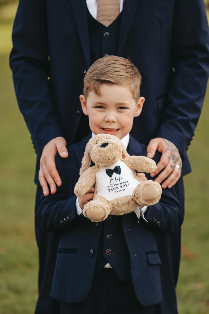In the scenic backdrop of Rudding Park, North Yorkshire, a young boy beams in a dark suit and tie, clutching a plush toy. His white shirt proudly declares "Best Dog and Ring Security." An adult in a matching ensemble stands behind him, hands reassuringly on his shoulders amidst the lush Harrogate greenery. © Aimee Lince Photography - Wedding photographer in Lincolnshire, Yorkshire & Nottinghamshire