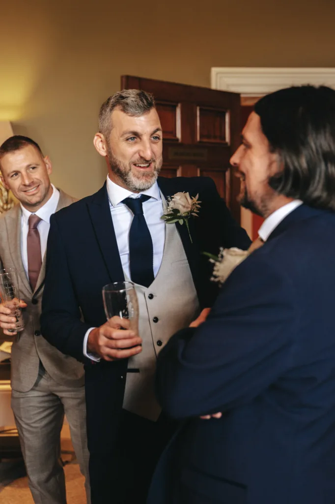 In a warmly-lit room at Rudding Park, three men in suits engage in friendly conversation. The central figure holds a drink, sporting a navy suit with a light gray waistcoat and a flower in his lapel. Another man, also adorned with a flower, listens intently as the third stands nearby with his drink. © Aimee Lince Photography - Wedding photographer in Lincolnshire, Yorkshire & Nottinghamshire