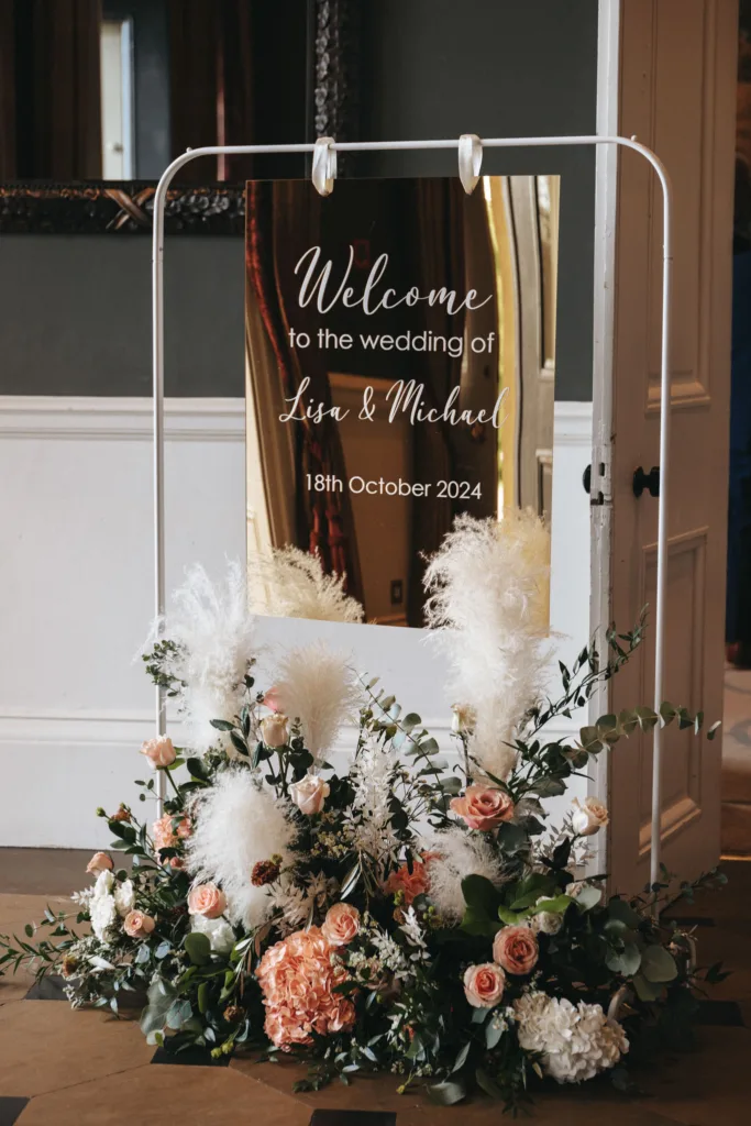 A wedding welcome sign with elegant script reading "Welcome to the wedding of Lisa & Michael, 18th October 2024" graces a white stand. A lush arrangement of white pampas grass, pink roses, and greenery decorates the base. Set indoors at Rudding Park with a glimpse of a door and mirror. © Aimee Lince Photography - Wedding photographer in Lincolnshire, Yorkshire & Nottinghamshire