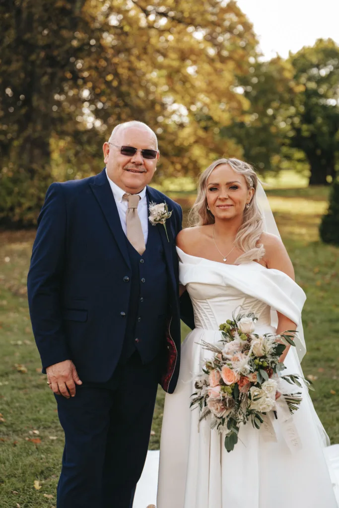 A smiling bride in an off-shoulder white dress holds a bouquet of white and peach flowers, standing next to a man in a dark blue suit with a beige tie and sunglasses. They are outdoors at Rudding Park, North Yorkshire, with grassy fields and trees painting the perfect Harrogate backdrop. © Aimee Lince Photography - Wedding photographer in Lincolnshire, Yorkshire & Nottinghamshire