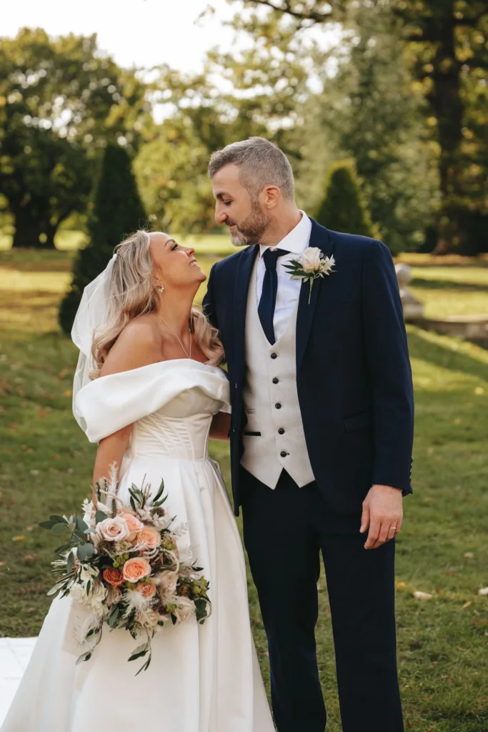 Bride and groom standing outdoors in a grassy area at Rudding Park, a picturesque spot in North Yorkshire. The bride wears an off-shoulder white gown and veil, holding a bouquet of pink and white flowers. The groom is in a dark suit with a white vest and navy tie. They gaze at each other, smiling warmly. © Aimee Lince Photography - Wedding photographer in Lincolnshire, Yorkshire & Nottinghamshire