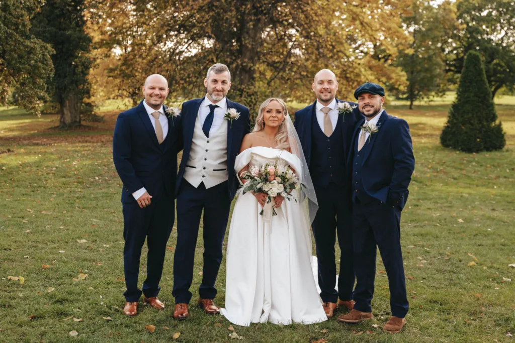 A bride in a white dress and veil stands with four men in navy suits and brown shoes on a grassy field at Rudding Park, Harrogate. The men wear ties or bow ties. The group is smiling against a backdrop of trees and the golden autumn foliage of North Yorkshire. The bride holds a bouquet of flowers. © Aimee Lince Photography - Wedding photographer in Lincolnshire, Yorkshire & Nottinghamshire