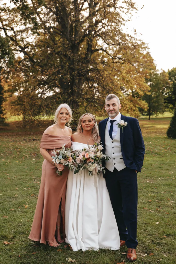 A bride stands between two companions, holding a bouquet. She wears a flowing off-shoulder white gown, while the woman beside her is in a pink dress and the man in a dark suit with a beige vest. They pose on lush grass at Rudding Park in Harrogate, with a large tree showing autumn colors behind them. © Aimee Lince Photography - Wedding photographer in Lincolnshire, Yorkshire & Nottinghamshire