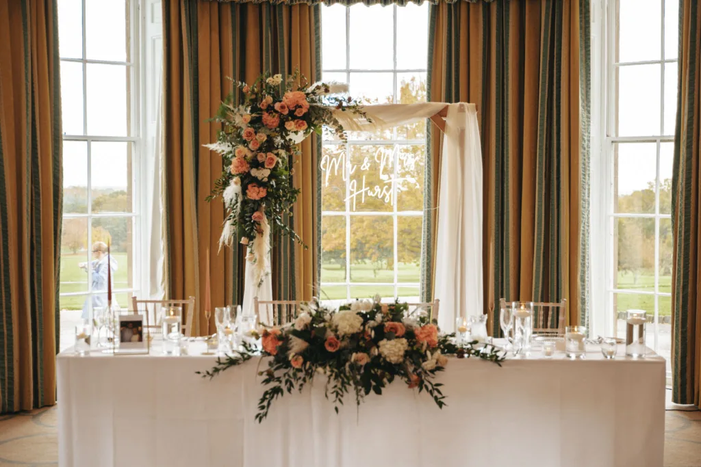 A wedding reception table is elegantly set with white linens and glassware at Rudding Park in Harrogate. A floral arrangement with pink and white flowers decorates the table and a nearby arch. Large windows offer a view of North Yorkshire's green lawn, enhancing the romantic and sophisticated atmosphere. © Aimee Lince Photography - Wedding photographer in Lincolnshire, Yorkshire & Nottinghamshire