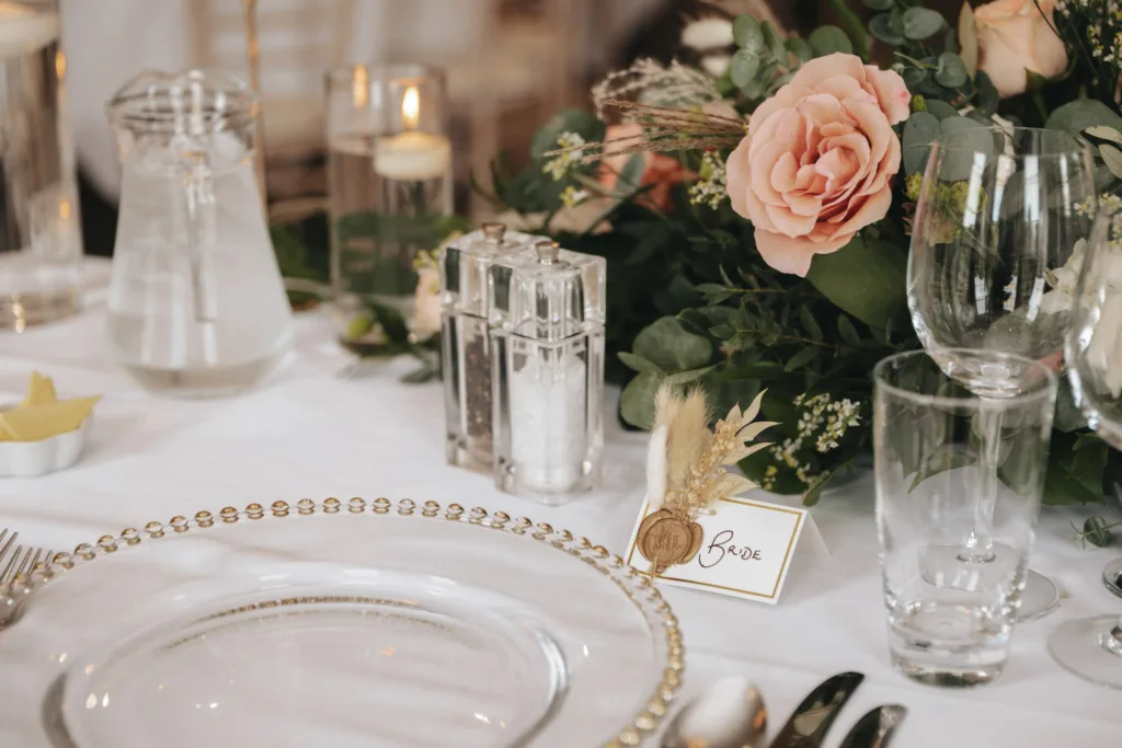 Elegant table setting at Rudding Park, Harrogate, featuring a clear charger plate trimmed with gold beads. Nearby are pepper and salt shakers, a candle, a water pitcher, glasses, and cutlery. A floral arrangement with pink roses and greenery decorates the table. A name card reads "Rose," adorned with wheat and rustic charm. © Aimee Lince Photography - Wedding photographer in Lincolnshire, Yorkshire & Nottinghamshire