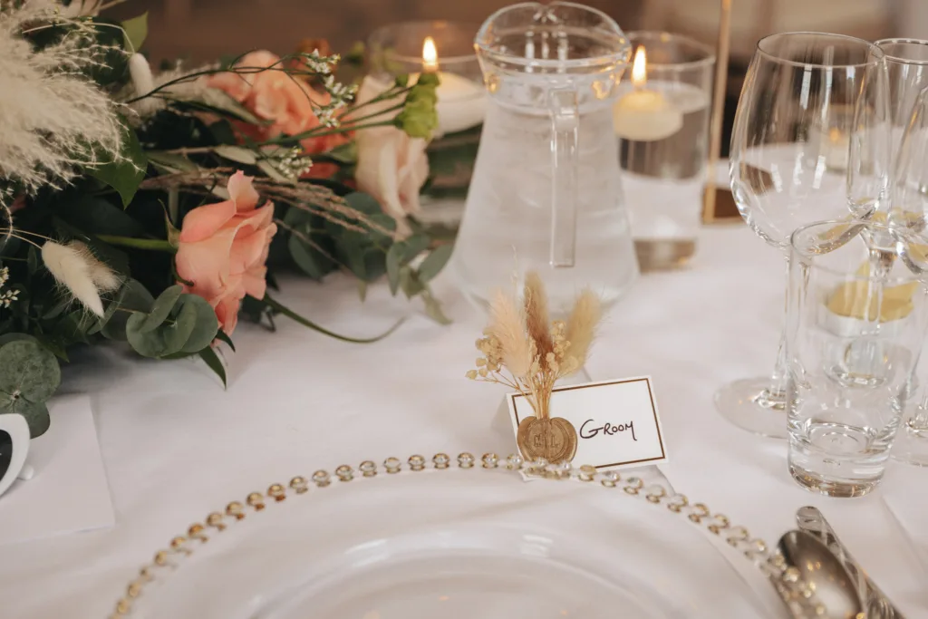 Elegant wedding table setting at Rudding Park in North Yorkshire features a clear glass plate on a beaded charger, wine glasses, and a water jug. A "Groom" place card with dried floral decor is displayed. Pink roses and candles enhance the ambiance on a white tablecloth in Harrogate. © Aimee Lince Photography - Wedding photographer in Lincolnshire, Yorkshire & Nottinghamshire