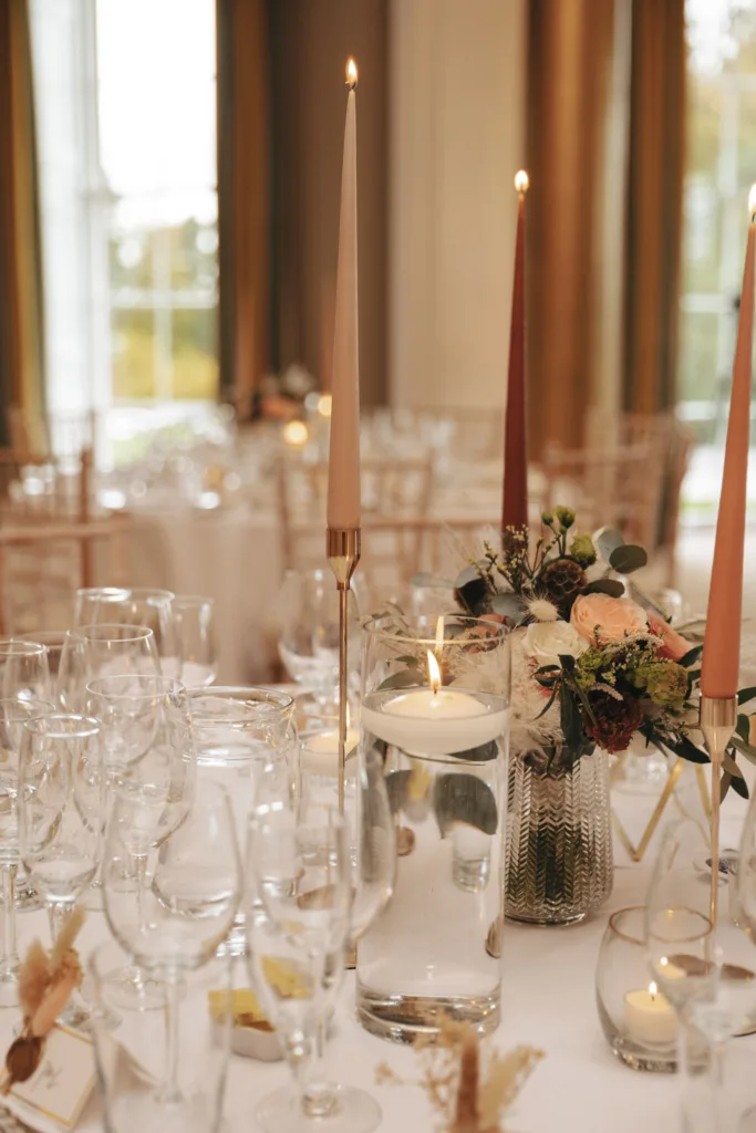 Elegant table setting at Rudding Park in North Yorkshire features tall, slender candles and a floating candle in a glass vase filled with water. A floral centerpiece with roses and greenery graces the table, while wine glasses and gold accents gleam softly under Harrogate's natural light filtering through grand windows. © Aimee Lince Photography - Wedding photographer in Lincolnshire, Yorkshire & Nottinghamshire