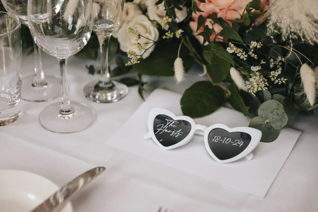 Heart-shaped sunglasses with "The Hursts" and "18-10-24" rest on a white envelope amid the refined elegance of Rudding Park. Nearby, wine glasses and a floral arrangement featuring white and peach blooms add charm to this Harrogate setting in North Yorkshire. © Aimee Lince Photography - Wedding photographer in Lincolnshire, Yorkshire & Nottinghamshire