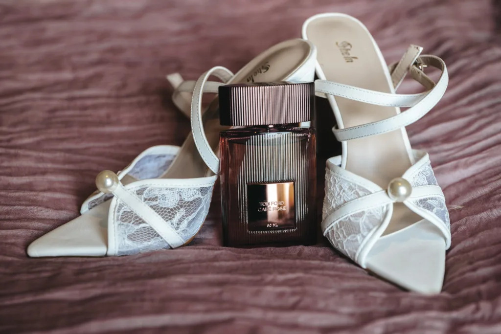 Elegant white lace and pearl high-heeled shoes rest on soft, textured mauve fabric, evoking the classic charm of Harrogate. Between them stands an upright rectangular bottle of Tom Ford perfume with a dark, glossy finish and metallic accents. The open-toe shoes boast a strappy design. © Aimee Lince Photography - Wedding photographer in Lincolnshire, Yorkshire & Nottinghamshire