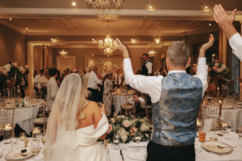 A bride and groom stand with their backs to the camera, facing a room full of cheering guests at a wedding reception in North Yorkshire. The bride is in an off-shoulder white gown, while the groom sports a blue patterned vest. Rudding Park's elegant floral arrangements and chandeliers grace the scene. © Aimee Lince Photography - Wedding photographer in Lincolnshire, Yorkshire & Nottinghamshire