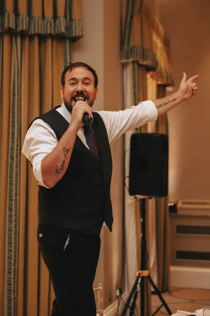 At a formal event in Harrogate, a bearded man is passionately speaking into a microphone with one hand raised. Dressed in a white shirt, black waistcoat, and tie, his tattooed arms add flair against the backdrop of yellow curtains and a speaker at what appears to be Rudding Park. © Aimee Lince Photography - Wedding photographer in Lincolnshire, Yorkshire & Nottinghamshire