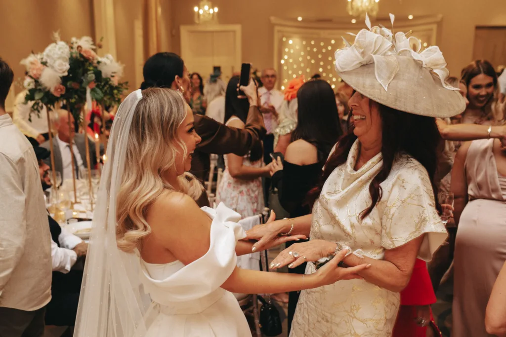 At a lively wedding reception at Rudding Park in Harrogate, a bride in a white dress and veil joyfully embraces a woman in a cream floral dress and matching hat. Guests in elegant attire celebrate around them, with floral arrangements and chandeliers visible in the warmly lit North Yorkshire room. © Aimee Lince Photography - Wedding photographer in Lincolnshire, Yorkshire & Nottinghamshire