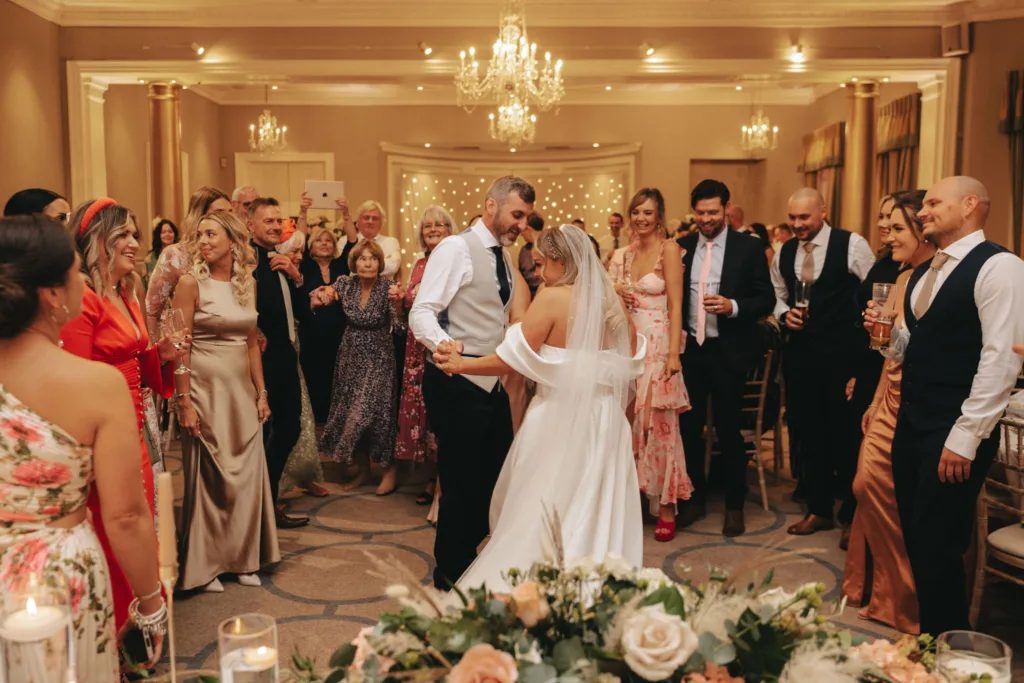 In a warmly lit reception hall at Rudding Park in North Yorkshire, a bride and groom share a dance. The bride wears a flowing white gown, the groom in a vest and tie. Guests smile and capture the moment as floral arrangements and chandeliers add to the festive ambiance in Harrogate. © Aimee Lince Photography - Wedding photographer in Lincolnshire, Yorkshire & Nottinghamshire