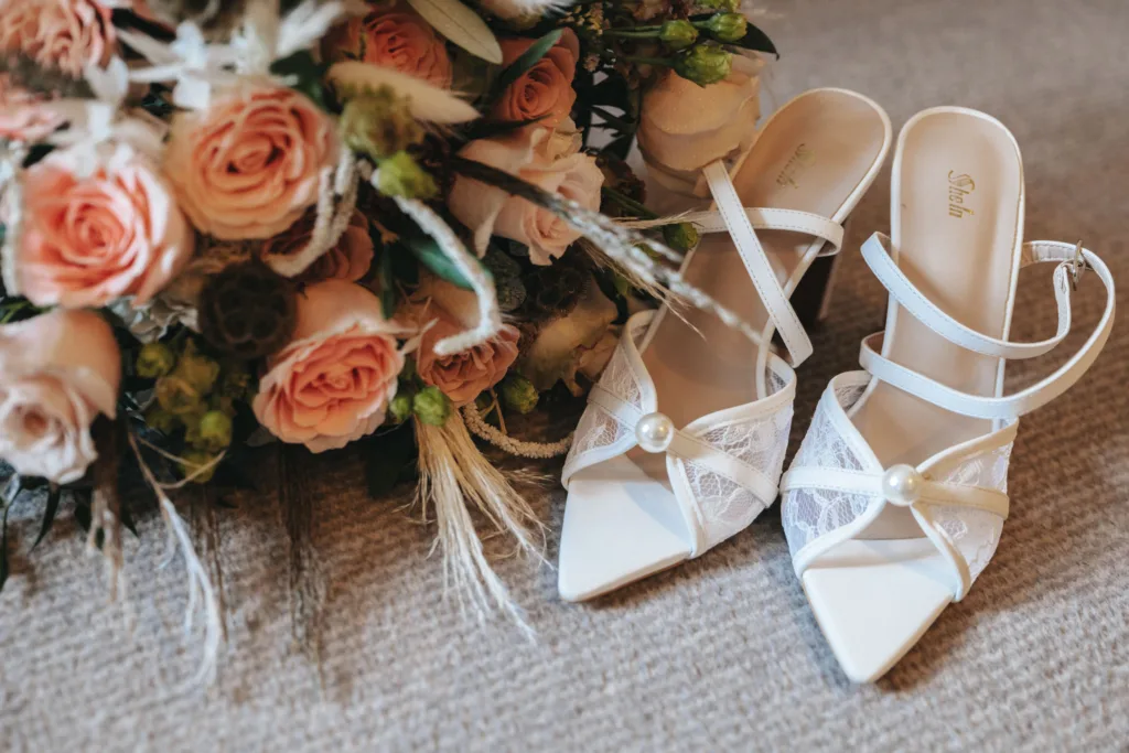 White strappy high-heeled shoes with pearl accents rest on a textured beige carpet next to a bouquet of pink roses and greenery. Evoking elegance and romance, these lace-detailed shoes complement a setting akin to a refined event in Harrogate, North Yorkshire. © Aimee Lince Photography - Wedding photographer in Lincolnshire, Yorkshire & Nottinghamshire
