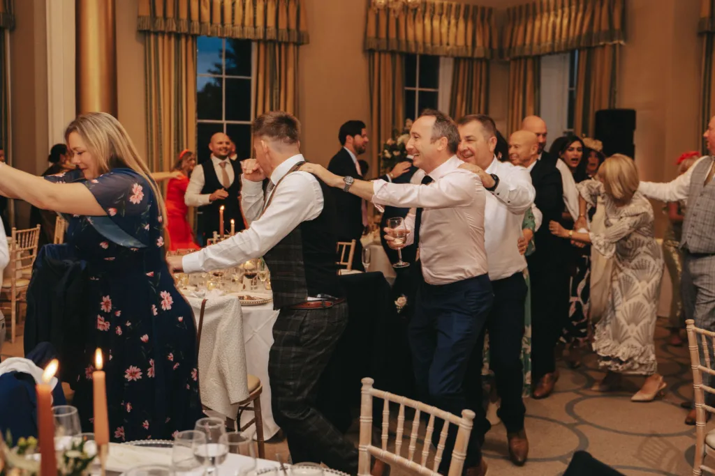 A joyful group of people are dancing in a conga line at an indoor event in Harrogate. Dressed in formal attire, with women in dresses and men in suits, they enjoy the celebration. The elegantly decorated room at Rudding Park features round tables, candles, and large windows highlighting North Yorkshire's charm. © Aimee Lince Photography - Wedding photographer in Lincolnshire, Yorkshire & Nottinghamshire