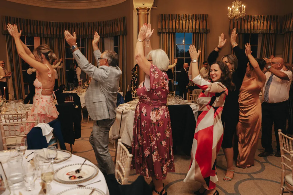 Guests at a formal event in Harrogate joyfully dance in a line, raising their arms. The women wear floral and colorful dresses, while the men are in suits. Round tables with white tablecloths and empty glasses are visible. Elegant drapes and a chandelier enhance the ambiance at Rudding Park. © Aimee Lince Photography - Wedding photographer in Lincolnshire, Yorkshire & Nottinghamshire