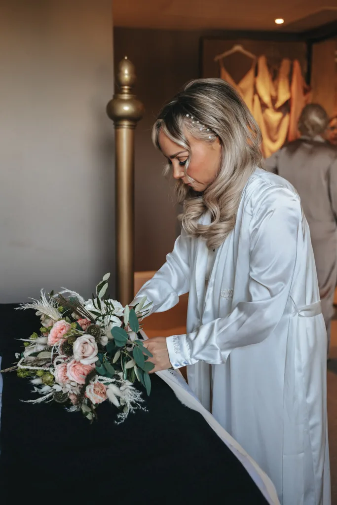 In a warmly lit room reminiscent of Rudding Park in Harrogate, North Yorkshire, a woman with long blonde hair in a white satin robe arranges pink and white flowers on a dark surface. A bronze pillar and blurred fabric add to the serene ambiance as another robed figure stands partially visible in the background. © Aimee Lince Photography - Wedding photographer in Lincolnshire, Yorkshire & Nottinghamshire